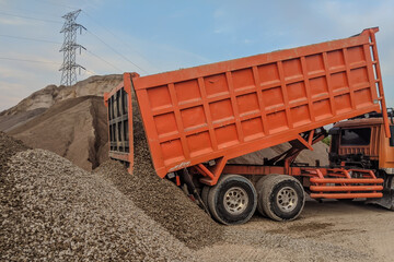 An orange dump truck unloads a pile of gravel at a construction or mining site, with a power line tower in the background. Illustrates heavy industry, earthmoving, and material transport.