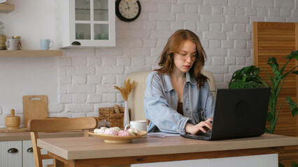 Engrossed in her work, a woman writes in a notebook while using a laptop in a bright kitchen. This image reflects the flexibility and comfort of remote work setups in modern homes.