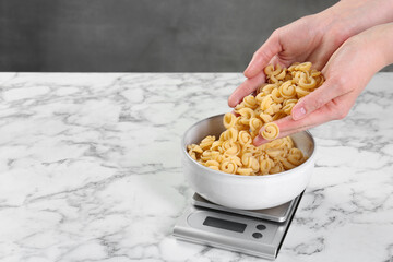 Woman adding pasta into bowl on kitchen scale on white marble table, closeup. Space for text