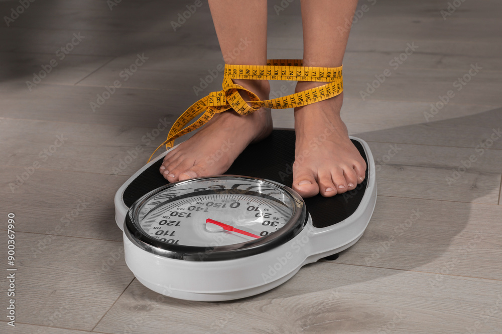 Poster Eating disorder. Woman tied with measuring tape standing on floor scale indoors, closeup