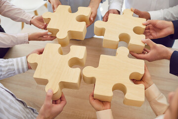 Close-up cropped shot of company employees hands holding fragments of wooden puzzle. Coworkers join...