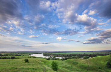 A verdant landscape unfolds under a sky adorned with fluffy white clouds, revealing a gentle river...
