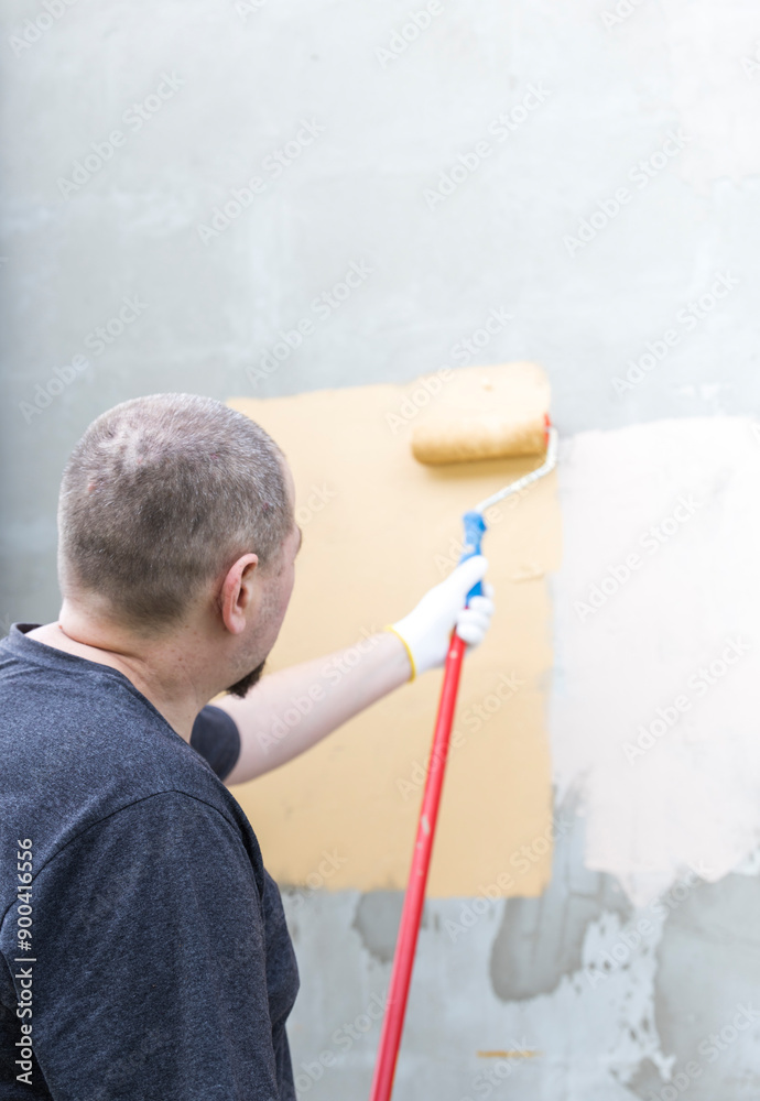 Wall mural a man is painting an exterior wall with a paint roller, applying a fresh coat of light yellow paint.