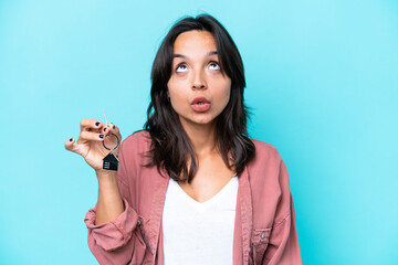 Young hispanic woman holding home keys isolated on blue background looking up and with surprised expression