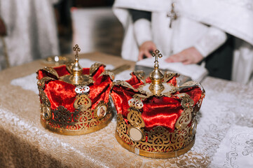 Two beautiful Christian crowns made of red fabric, with a gold frame, a pattern, and a cross lie on a table with a tablecloth at a wedding ceremony, a rite in the church. Photography, religion.