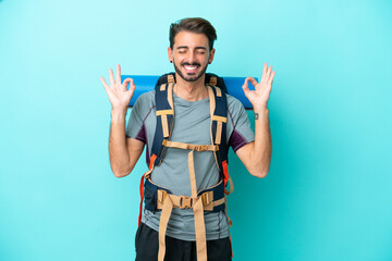 Young mountaineer man with a big backpack isolated on blue background in zen pose