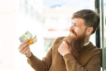 Redhead man with beard taking a lot of money at outdoors thinking an idea and looking side