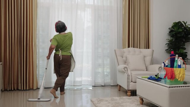 Handheld Shot Footage A housekeeper in a green apron and red headphones thoroughly mops the living room floor, singing and dancing in spotless and bright environment.