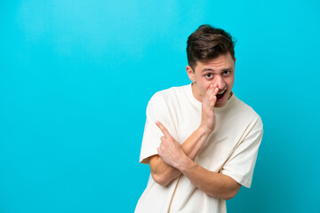 Young handsome Brazilian man isolated on blue background pointing to the side to present a product and whispering something