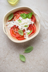 Sliced tomatoes, stracciatella cheese and green basil in a serving tray, vertical shot on a beige stone background, top view