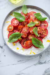 Plate of fresh stracciatella di bufala with red tomatoes and green basil, vertical shot on a white marble background, elevated view