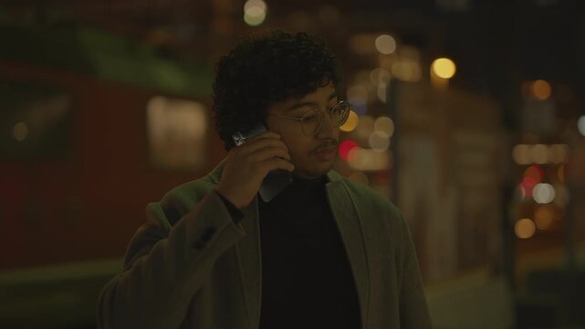 A man wearing glasses in a cityscape at night, lost in thought while holding a smartphone
