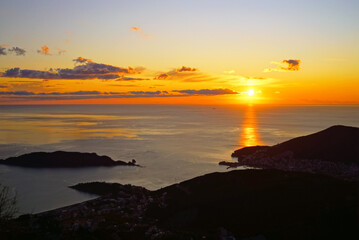Sunset over Budva (Montenegro): view of the coast and the island of St. Nicholas in the evening. The sun setting behind the sea, bright colors of the sunset sky with light clouds and the Adriatic Sea