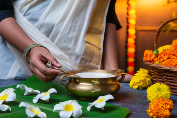Onam festival sweet dessert Pal payasam, Woman wearing traditional saree and stirring payasam in uruli and flower decoration on the background