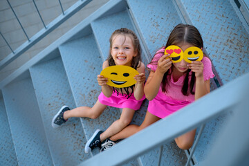 A funny girl holds a winking cardboard yellow emoji. The girl closes her eyes with different emoticons