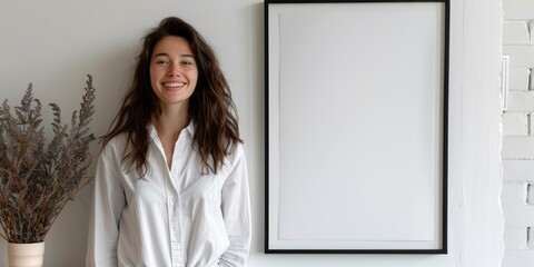 A smiling woman is standing next to a blank poster in a frame and decorating the wall on a white background in the living room.
