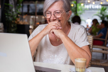 Elderly woman in video call while enjoying a coffee at a cafe. Video call by laptop for a cheerful senior female, highlighting happiness and contentment in relaxed atmosphere