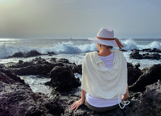 Back view of senior woman sitting on a rocky beach watching ocean waves crashing with white foam. Concept of relaxed elderly person admiring the strength of the sea