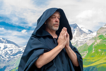 An elderly monk in a black hooded cassock meditates and prays among alpine mountains under a blue sky