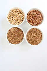 Four white bowls filled with chickpeas, kidney beans, lentils and buckwheat groats are arranged in the shape of a square on a white background
