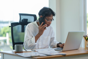 A professional man in a white shirt is working at his desk, engaged in a phone call while using his laptop. He is seated in a modern office.