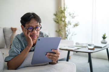 Young man is lounging on a sofa in a cozy living room, deeply engrossed in his tablet. He is wearing glasses and resting his head on one hand, appearing focused and thoughtful.