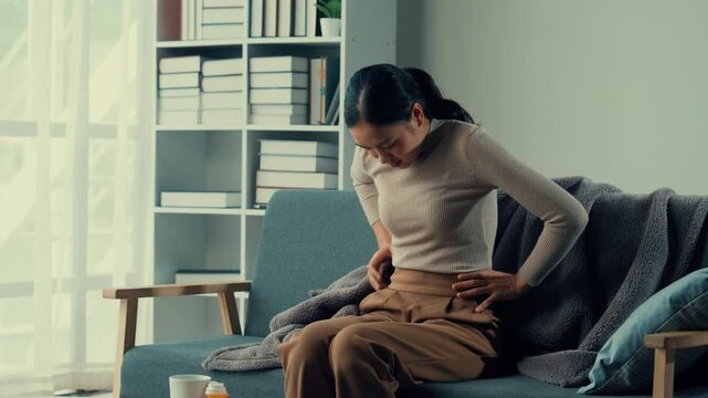 Female office worker at home suffering back pain on sofa, trying to find relief through stretching and adjusting posture