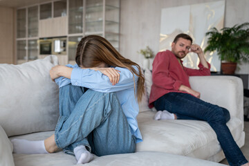 Woman in tears, sadness, hides face and shrinks on couch, while her distressed husband sits at...