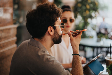 Young man recording a voice message on smart phone while sitting at an outdoor cafe with a female friend wearing sunglasses.