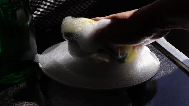 Un primer plano de una mano de mujer que presiona una esponja amarilla para lavar un plato de color blanco lleno de jab&oacute;n l&iacute;quido con espuma en la cocina
