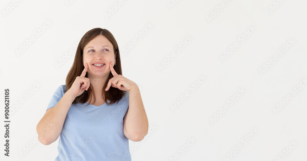 Wall mural Young brunette woman pointing fingers at her cheeks isolated over white background