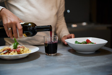 Person pouring red wine into a glass during a pasta dinner in a cozy home setting