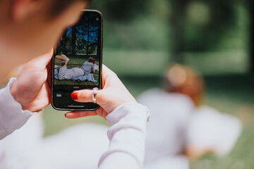 In a tranquil park setting, a woman uses her smart phone to capture a serene moment of her friend lounging on the grass, embodying relaxation and friendship.