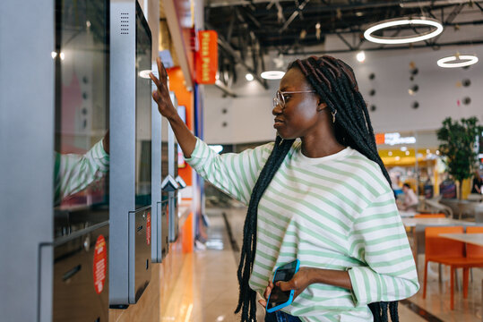 Black Woman with glasses and striped shirt using a touch screen kiosk in a modern shopping mall. Girl chooses burger on touch menu of fast food restaurant. 
