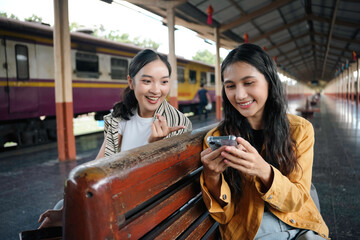 Two happy young women are sitting on a bench at a train station, using a smartphone and smiling