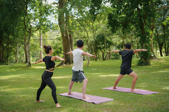 Three asian people are improving their balance and flexibility by practicing yoga warrior two pose on mats in the park