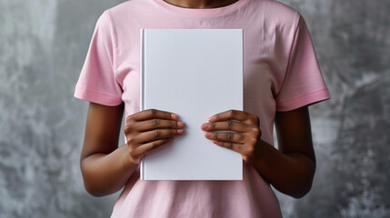 A person holding a blank white book in front of their chest, wearing a pink shirt.