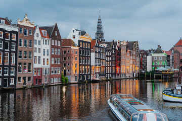 Beautiful Amsterdam canal with iconic traditional houses and boat at dusk, reflecting in the calm waters under a cloudy sky.