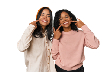 Young Latin friends together in studio smiles, pointing fingers at mouth.
