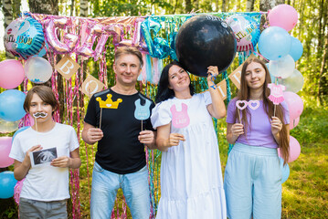 A family with two children at a baby gender reveal party. They hold a black balloon with the words...
