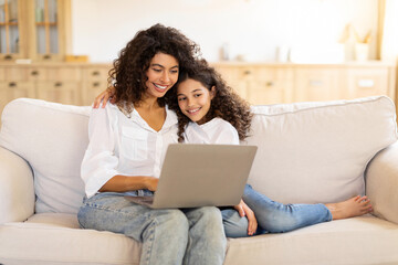 Happy young mother and daughter sitting on couch at home using laptop together, browsing internet on computer or choosing movie to watch