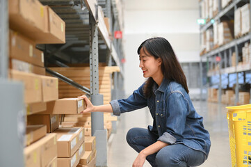 Woman squatting and selecting a box from a shelf in a warehouse, smiling and focused.
