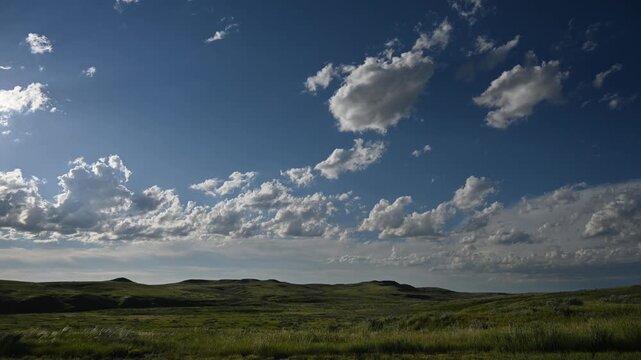 Time lapse of white summer clouds moving above a prairie badlands setting with blowing grass and distant rolling hills.  The sunlight hitting the scene varies as the clouds move.  
