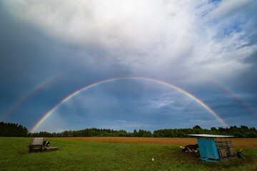 Two rainbows after a storm over a pasture, with cows, a watering hole, a feeding trough.
