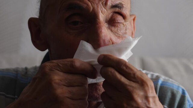 Caucasian pensioner 70-79 years old with a runny nose is unwell, wipes snot and coughs, an elderly person with a cold and fever coughs while inside