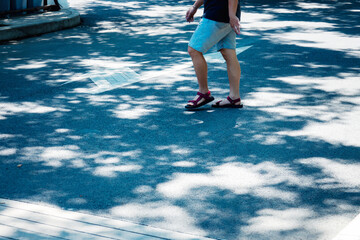 A man is walking on a sidewalk in the shade. He is wearing a blue shirt and gray shorts
