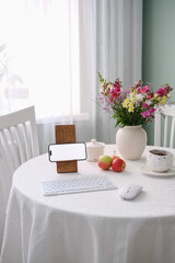 A cozy home office with a flowers in ceramic vase, apples, cup of tea, modern tech gadgets: mobile phone, keyboard, mouse.