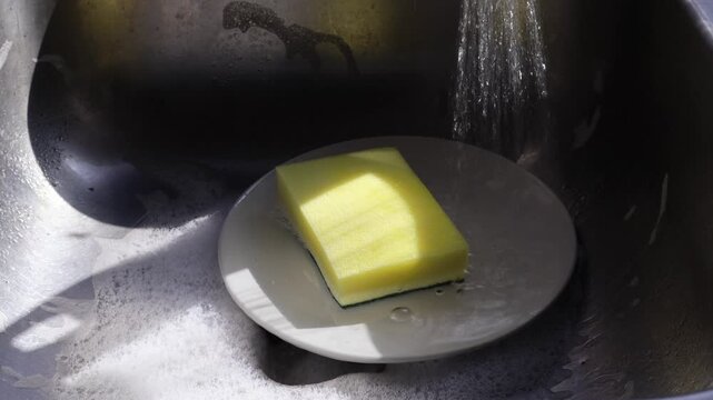 Un primer plano de una esponja amarilla llena de jab&oacute;n liquido que le cae agua en un plato de color blanco dentro de un fregadero