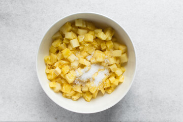 Overhead view of diced pineapple and sugar being mixed together in a white bowl, process of making pineapple cheong or pineapple syrup