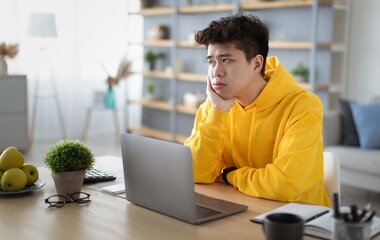 Bored young Asian guy sitting at desk using pc laptop, looking aside, thinking about something...